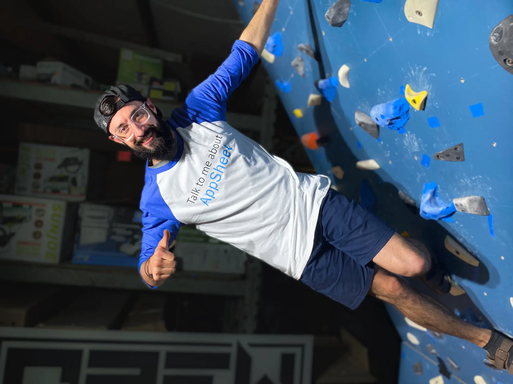 A team member hanging off an indoor climbing wall in a QREW t-shirt, grinning at the camera.