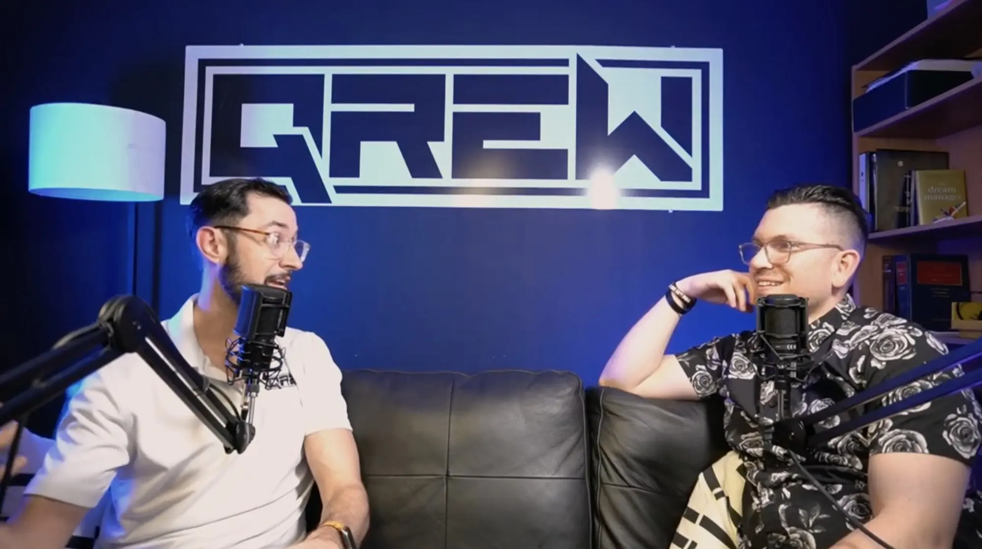 Two hosts recording a podcast on the QREW set, the illuminated QREW logo on the wall behind them.